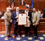 four men in the front of the Connecticut House of Representatives chamber. Three of them are wearing business clothes, while Mark Sheldon is wearing Revolutionary War costume and holding a copy of the Declaration of Independence.