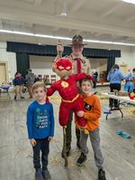 a scout and children posing with a superhero-themed scarecrow